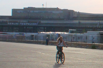 Au fond, les bâtiments de l'aéroport de Berlin-Tempelhof, au milieu le village de réfugiés, devant, une cycliste sur une ancienne piste d’atterrissage 